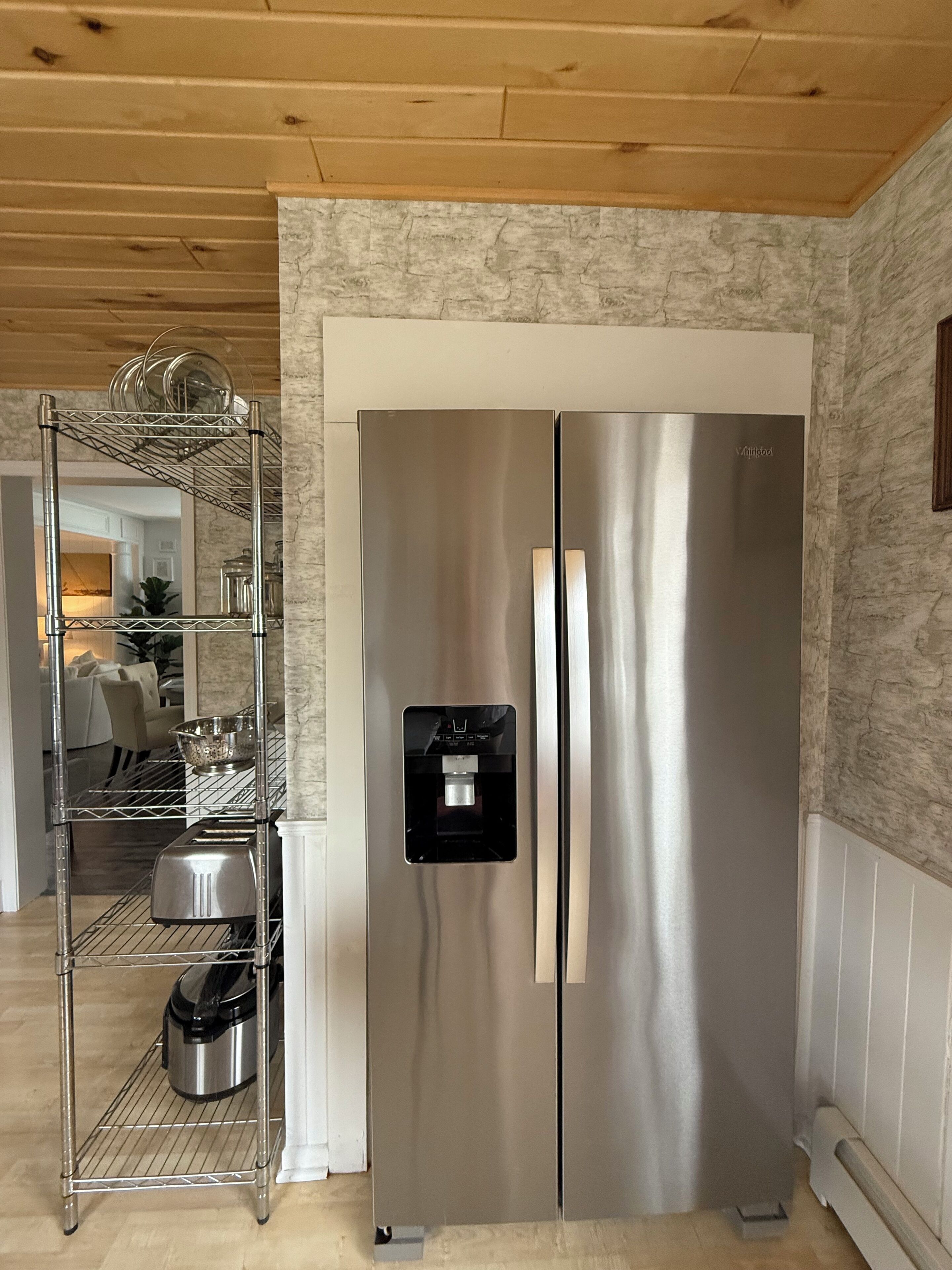 A stainless steel refrigerator with a water dispenser, a metal shelf unit with dishes, and a marble-patterned wall.