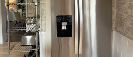 A stainless steel refrigerator with a water dispenser, a metal shelf unit with dishes, and a marble-patterned wall.
