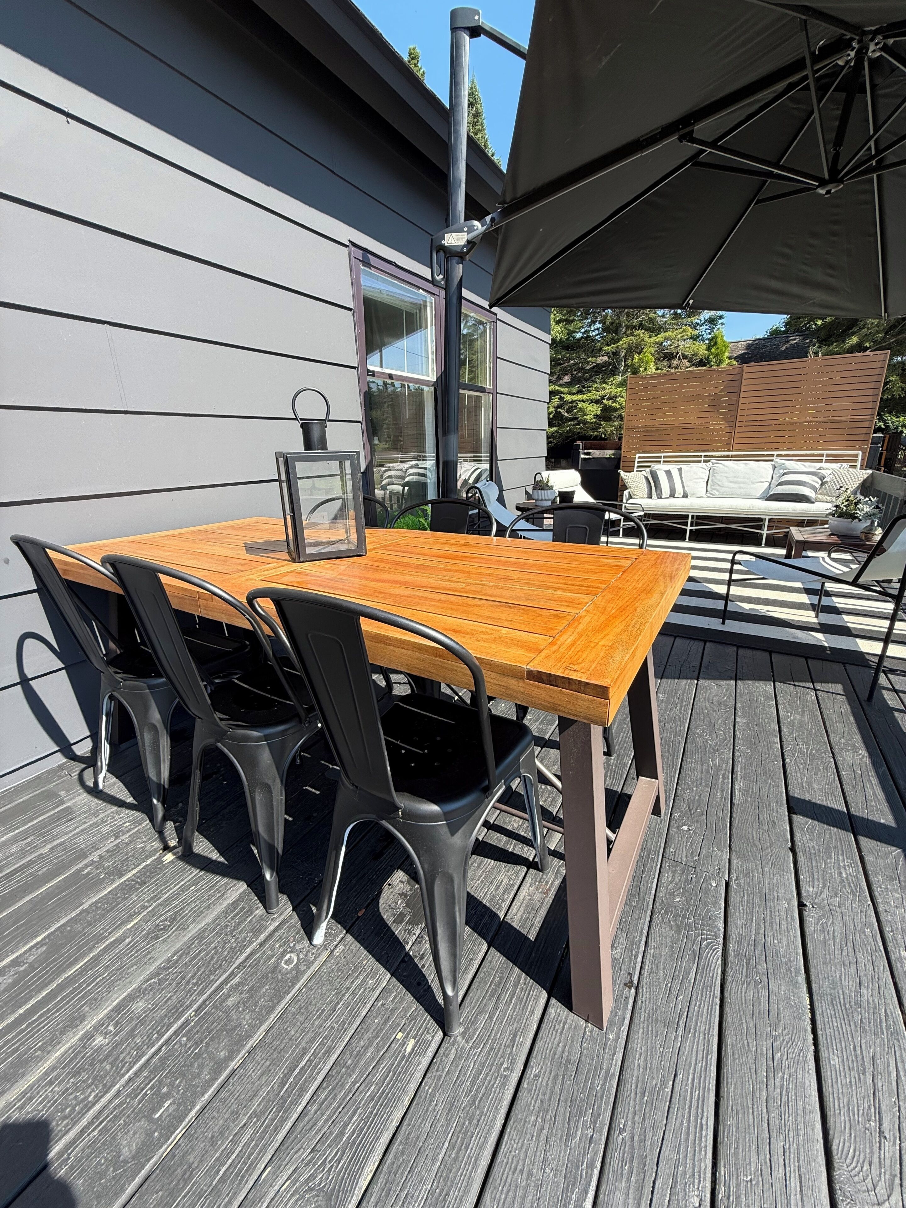 An outdoor patio with a wooden table and chairs, a hanging lantern, and a covered area with a sofa and a grill.