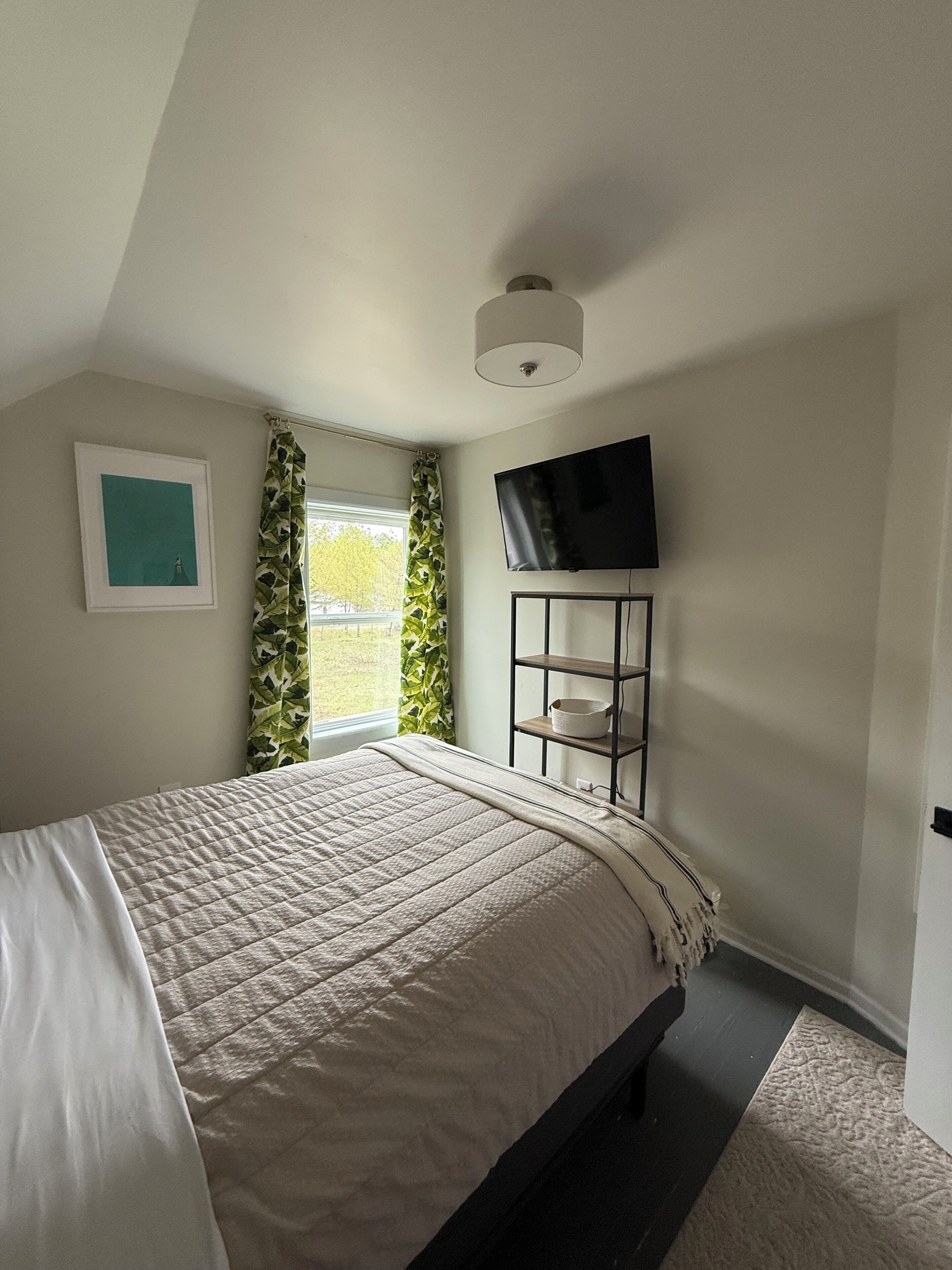 Un dormitorio con una cama, un televisor montado, un estante y una ventana con cortinas.