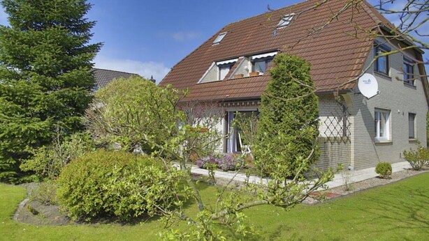 Une maison à deux étages, dotée d’un toit en pente, d’une antenne parabolique et d’un jardin bien entretenu, agrémenté de divers arbustes et d’une pelouse.
