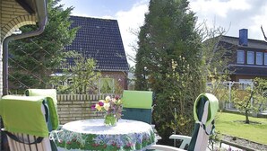 Une terrasse extérieure avec une table ronde et quatre chaises, entourée d’un jardin où poussent différentes plantes, et un mur de briques en arrière-plan.