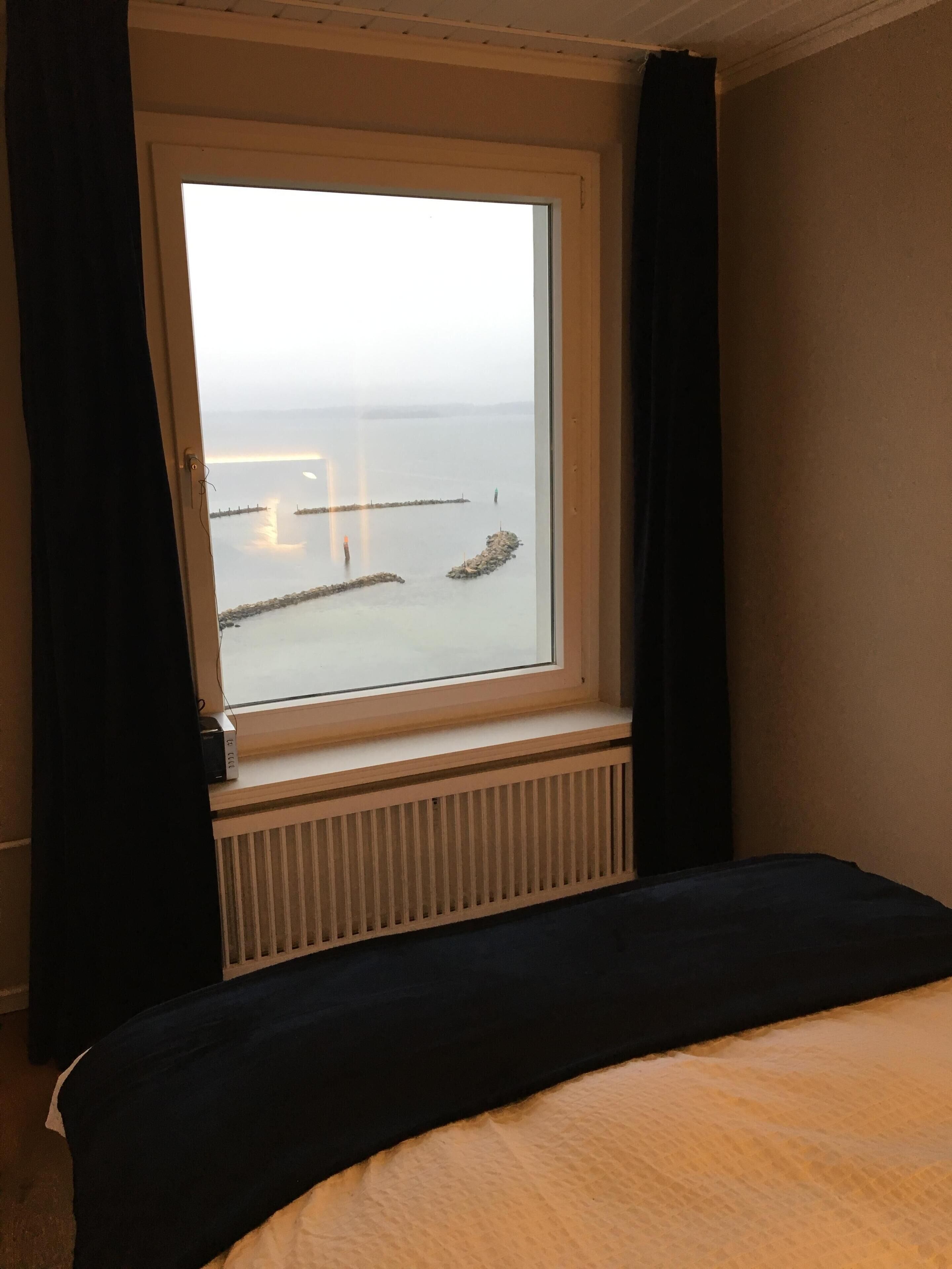 Habitación de hotel con un ventanal grande que da a una masa de agua tranquila, una cama y un radiador debajo de la ventana.