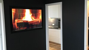 A modern living room with a fireplace, wooden flooring, and a kitchen visible through an open doorway.