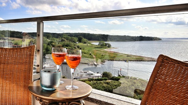 A table with two glasses of red wine and a metal container on a balcony overlooking a body of water and greenery.