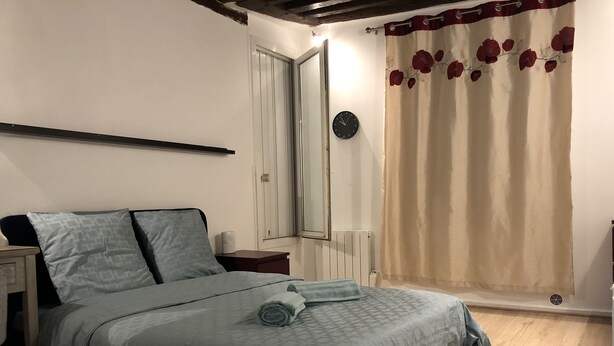 A bedroom with a bed, a nightstand, a mirror, a clock, and a window with curtains. - romantic paris in the heart of paris (Paris)