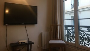 A room with a mounted television, a small table with a box, a radiator, and a window with a decorative iron railing. - romantic paris in the heart of paris (Paris)