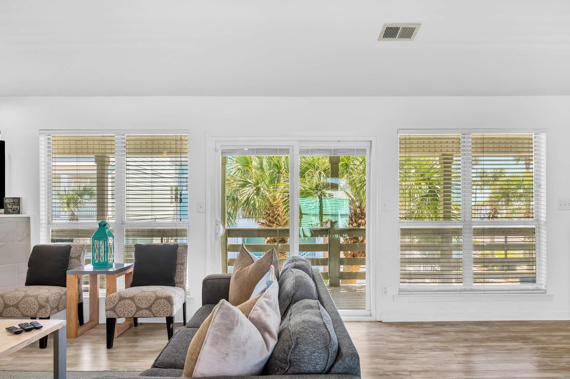 Uma sala de estar ampla com um sofá cinzento, poltronas estampadas e uma mesinha com controles remotos. Grandes janelas com persianas oferecem vista para palmeiras e uma construção verde lá fora.