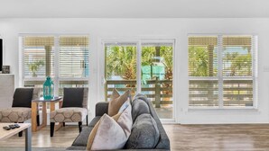 A spacious living room with a grey sofa, patterned armchairs, and a small table with remotes. Large windows with blinds offer views of palm trees and a green structure outside. - Gulf view, Steps to beach, heated pool, walk to restaurants (Destin)