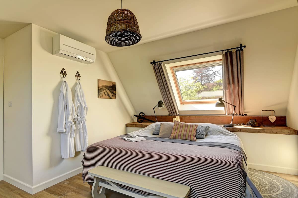 Un dormitorio con cama, un banco, una ventana con cortinas, una mesita de noche y un aire acondicionado de pared.