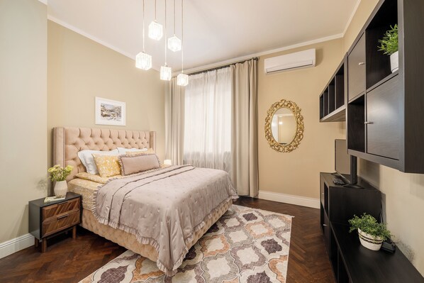 Un dormitorio con cabecera capitonada, cama con colcha estampada, mesita de noche con un florero, un espejo grande y estanterías empotradas.