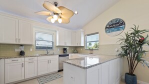 Une cuisine avec des meubles blancs, un plan de travail en granit, un ventilateur de plafond, une horloge et une plante en pot.