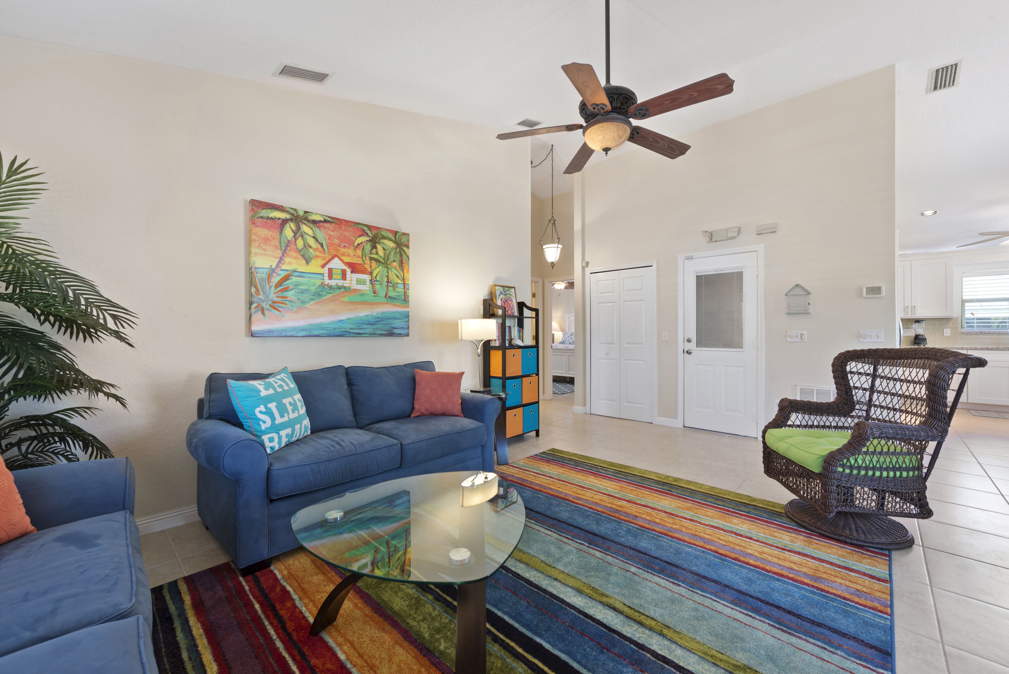 A living room with a blue sofa, a glass coffee table, a colorful striped rug, a wicker chair, and a ceiling fan.