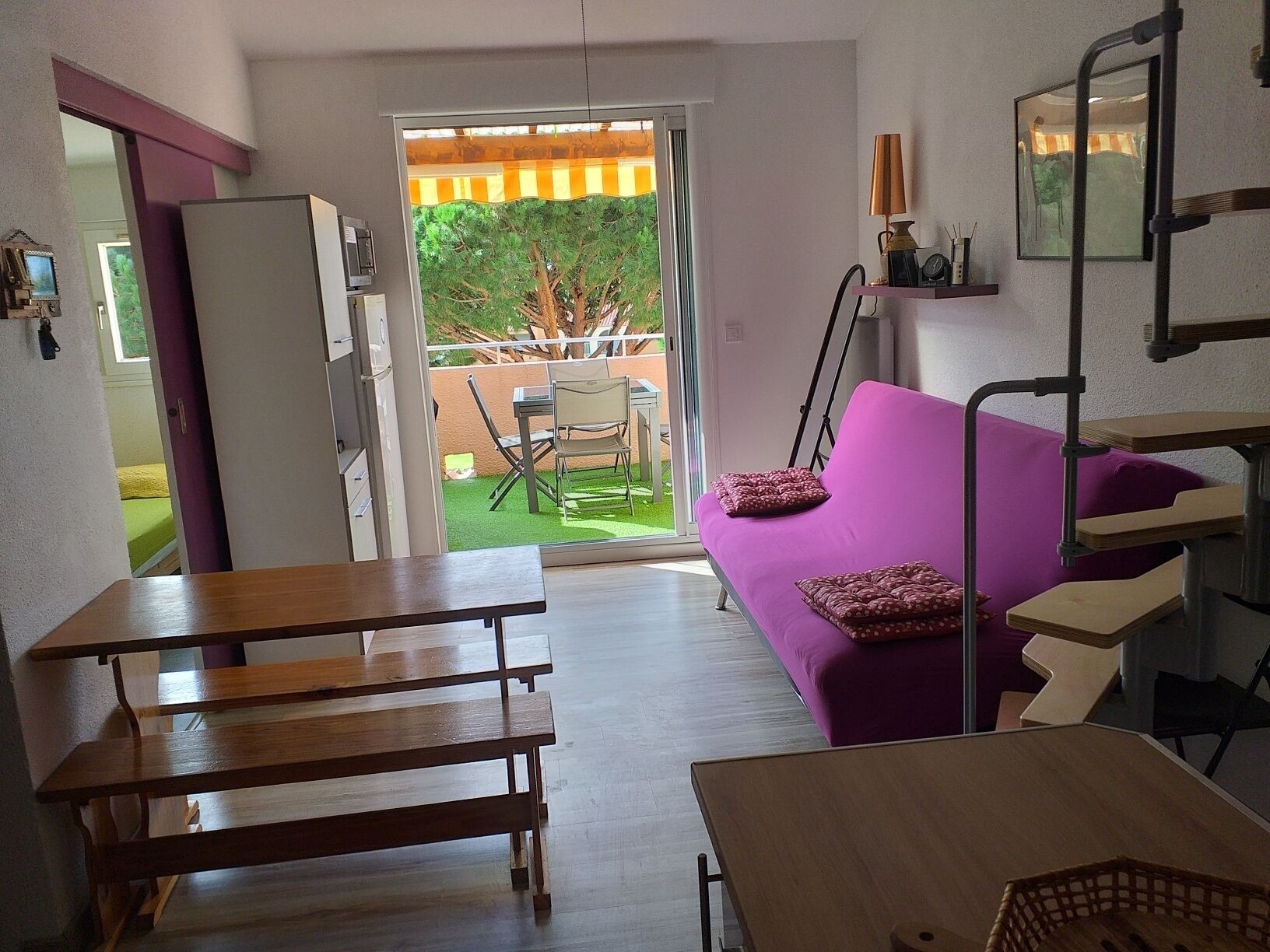 Un espace de vie compact avec un canapé violet, des tables en bois et une porte-fenêtre coulissante donnant sur un espace extérieur équipé d’une table et de chaises.