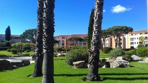 A courtyard with palm trees, a stone pathway, and a building with a red roof in the background. - RESIDENCE IN SEASIDE (La londe les maures)
