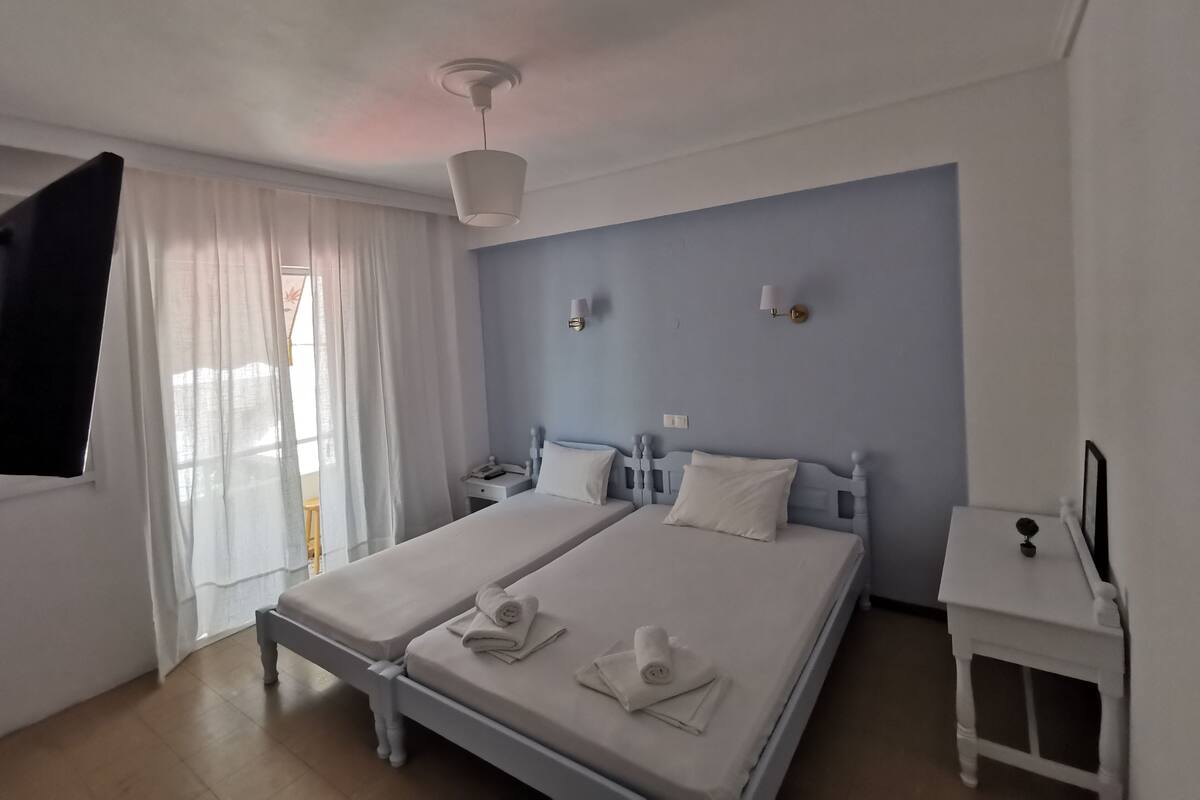 A hotel room with two beds, a bedside table, a nightstand, a lamp, a window with curtains, and a ceiling light.