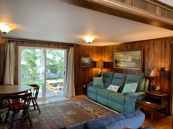 A cozy living room with a blue sofa, wooden walls, a dining table with chairs, and a view through the glass door.