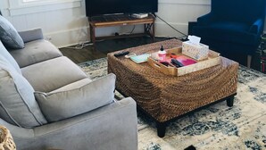 Sala de estar con sofá gris, sillón azul, televisión sobre un mueble de madera, reposapiés tejido y alfombra estampada.