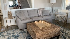 Una sala de estar con un sofá gris, un otoman tejido, una mesita redonda pequeña, una lámpara de pie y una escalera.