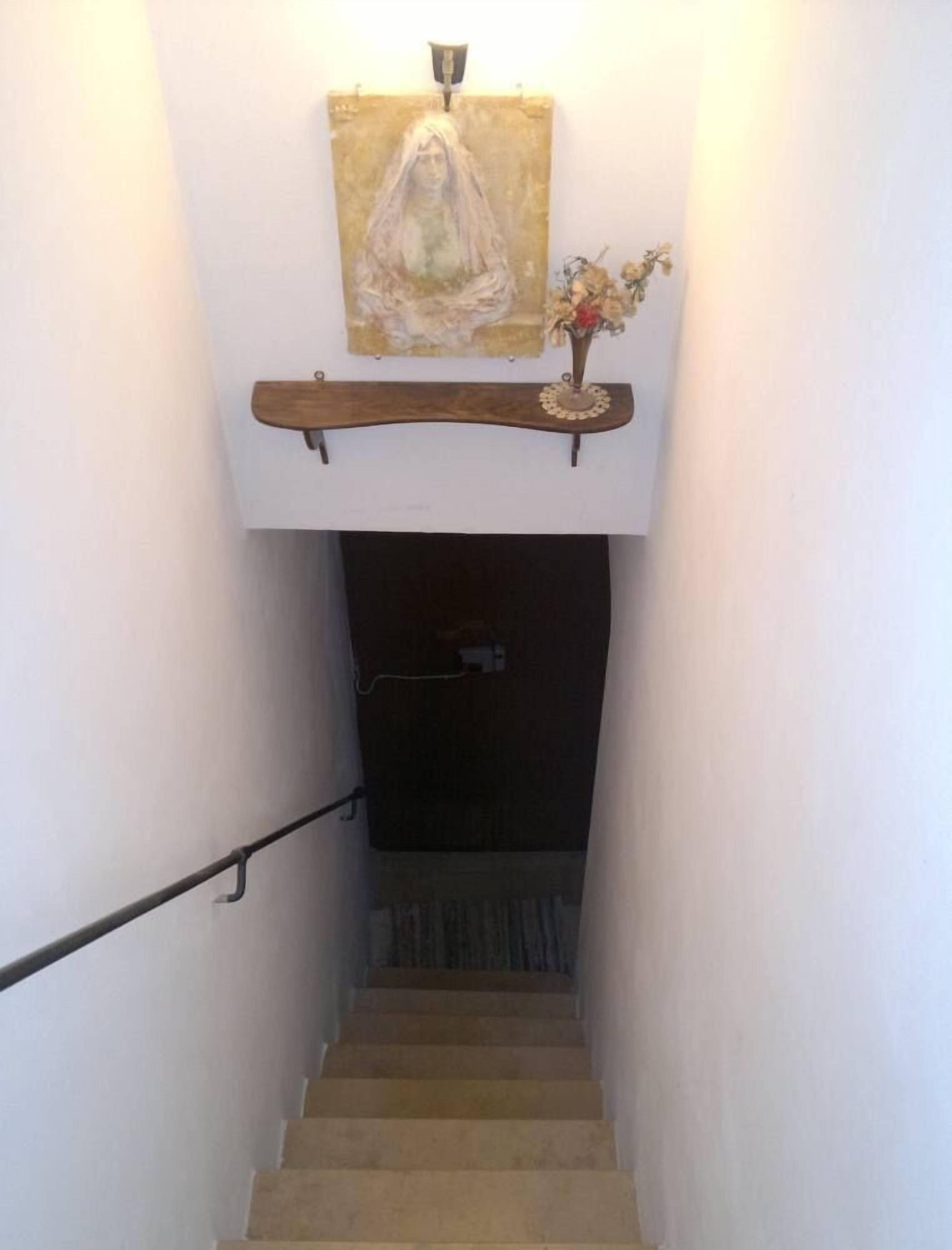 Un escalier avec une rampe en bois, une étagère fixée au mur sur laquelle est posé un vase de fleurs séchées, et un tableau au-dessus de l’étagère.