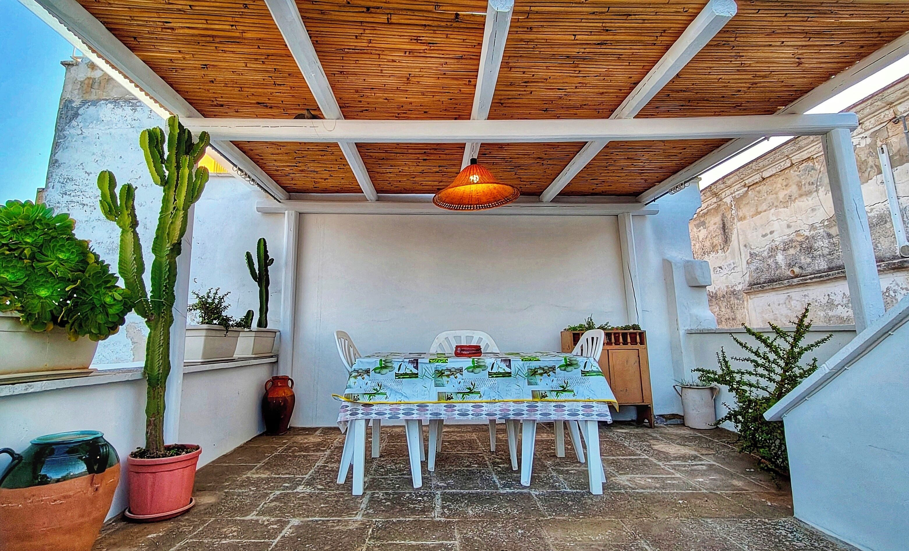 A covered patio with a table and chairs, surrounded by potted plants and a wooden ceiling.