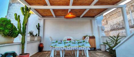 A covered patio with a table and chairs, surrounded by potted plants and a wooden ceiling.