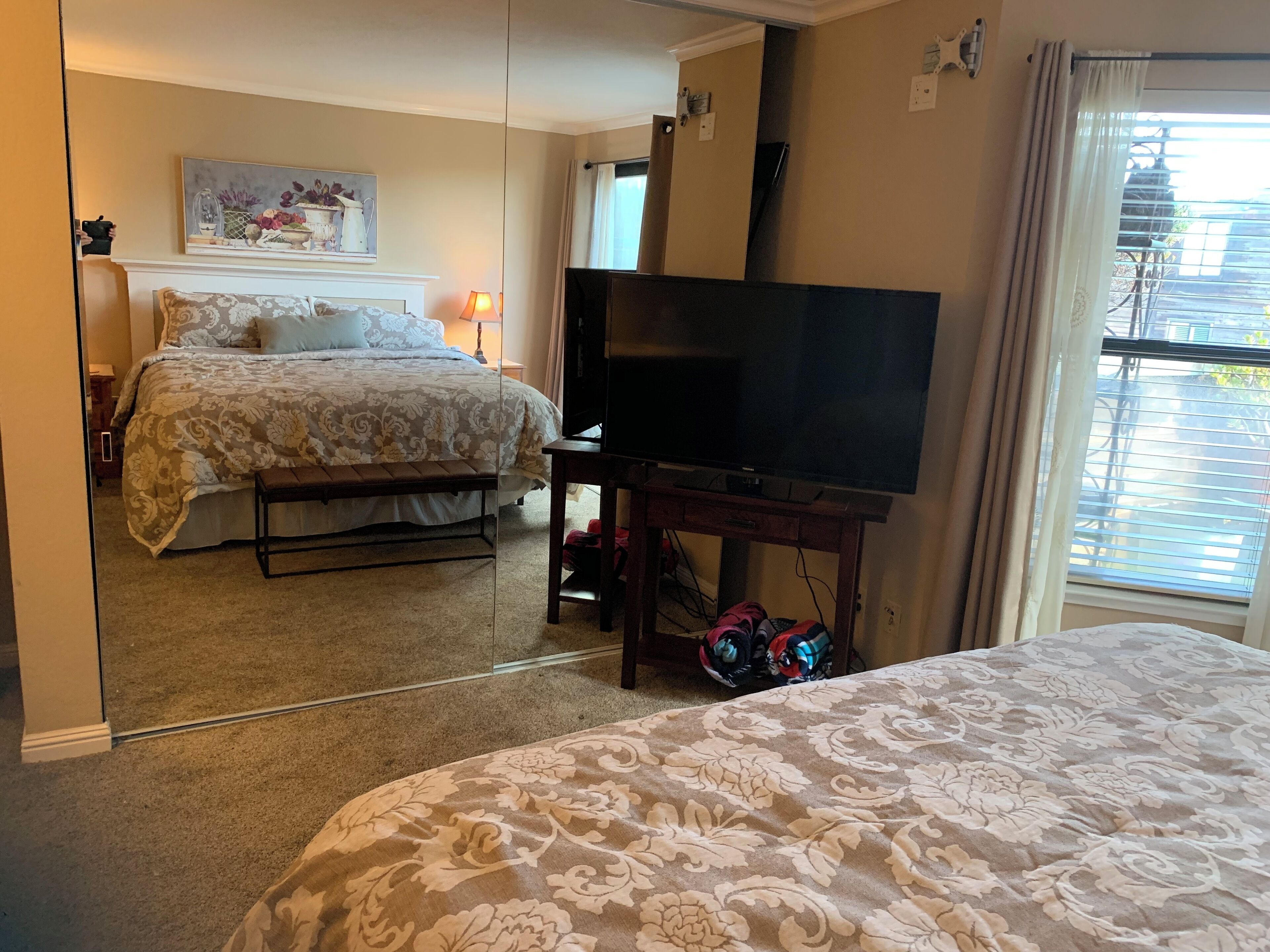 Une chambre à coucher avec un lit, un téléviseur posé sur un meuble et une fenêtre avec des stores.