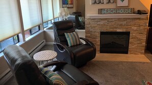 Un salon avec une cheminée, un canapé, un fauteuil inclinable et une petite table ronde. De grandes fenêtres sont équipées de stores, un tableau représentant des bouteilles est accroché au mur, et une enseigne indique « Beach House ».