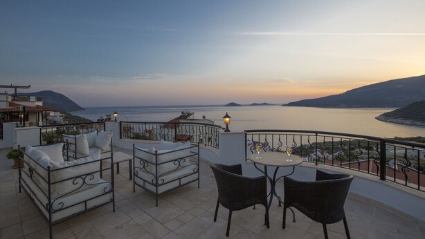 Eine Terrasse mit einem Bett, zwei Stühlen und einem Tisch mit einem Weinglas, mit Blick auf ein Gewässer und Hügel.