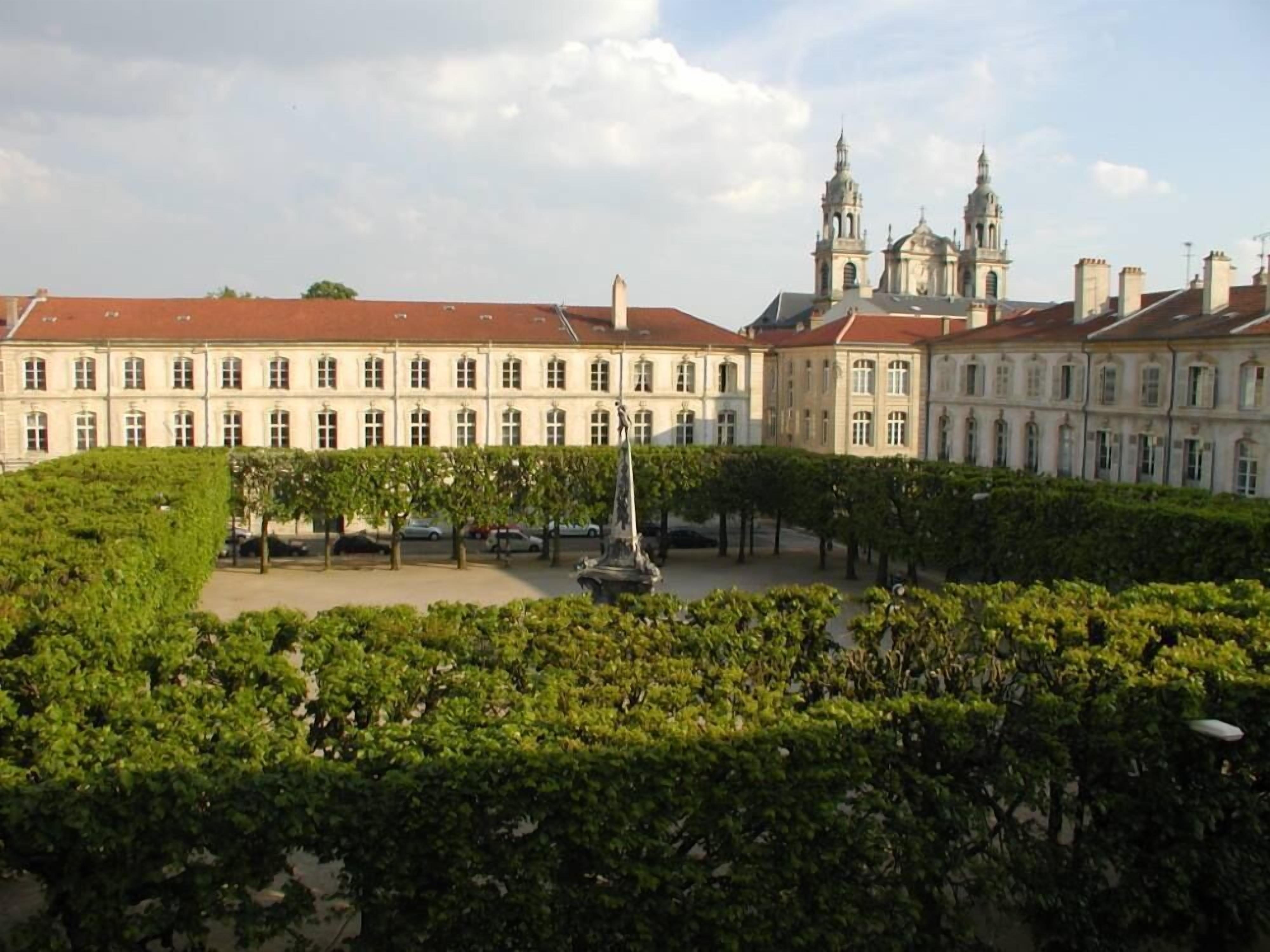 A large, historic building with a symmetrical facade, a central courtyard with a statue, and a church with twin spires in the background.
