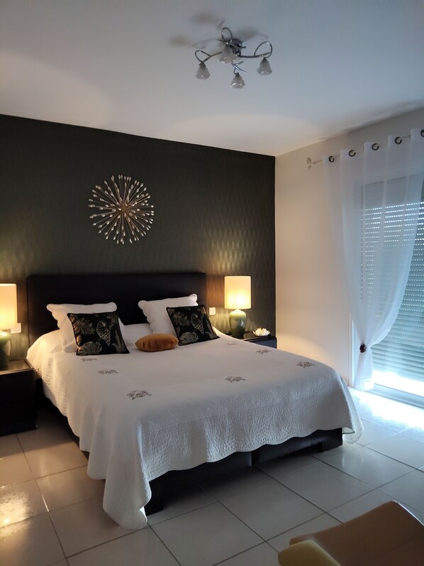 Une chambre bien éclairée, avec un grand lit, deux tables de chevet avec des lampes, un élément décoratif mural et une fenêtre avec des rideaux.