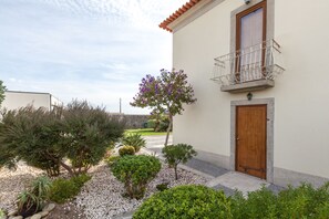 Um edifício com porta de madeira, varanda com grade de metal e jardim com diversas plantas e uma árvore com flores roxas.