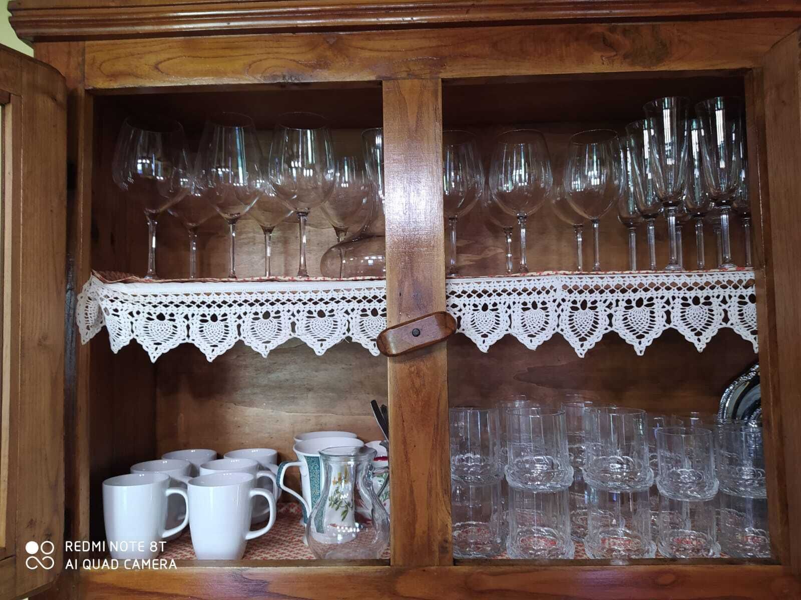 Un armario de madera con dos estantes, uno con copas de vino y el otro con tazas y una jarra.