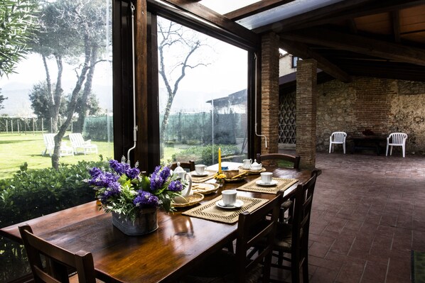 A wooden dining table set with white chairs, a vase of purple flowers, and a brick wall in the background.