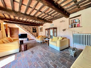 Una sala de estar acogedora con suelo de piedra, vigas de madera, una escalera, televisor y estantería.