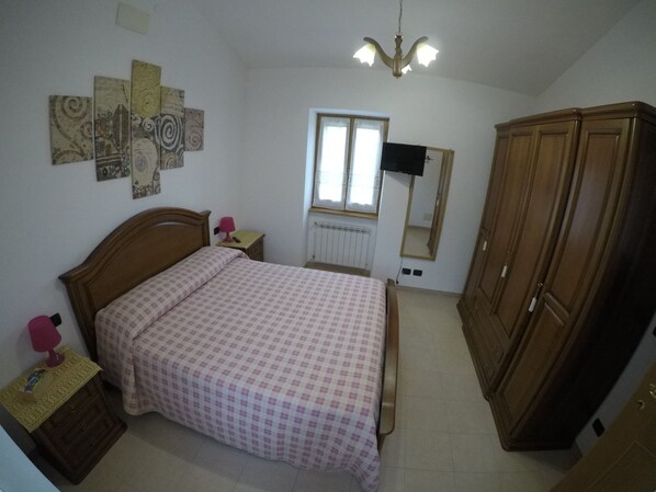 A bedroom with a bed, wooden wardrobe, a nightstand, a lamp, a wall-mounted TV, and a window with curtains. - Roccascalegna, Chieti (Roccascalegna)