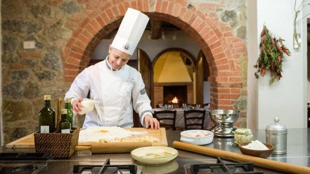 Un chef con uniforme blanco y gorro alto, preparando comida en una barra con diversos ingredientes y utensilios de cocina.