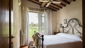 Un dormitorio con cama, ventilador de techo, ventana con cortinas y vistas a un jardín.