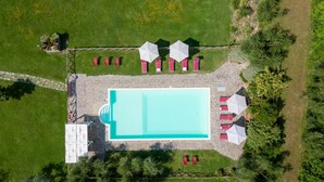 Vista aérea de una piscina rectangular rodeada de sillas de descanso rojas y sombrillas blancas, con un muro de piedra y follaje circundante.