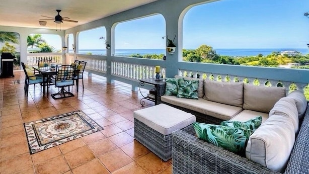 A spacious balcony with a view of the ocean, a dining area with chairs and a table, and a seating area with a sofa and cushions. - Mountaintop Villa with Spectacular Sunset View of Caribbean  (rincon)