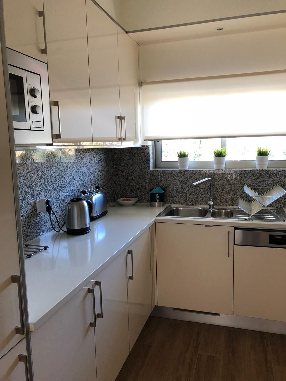 A modern kitchen with white cabinets, granite countertops, a built-in oven, a kettle, a sink, and a window with potted plants.