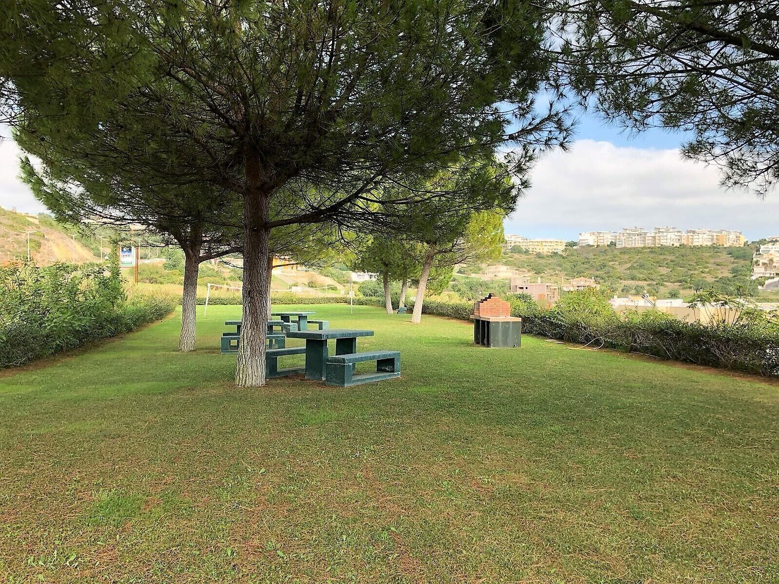 Un espacio al aire libre con césped, mesas de picnic y una pequeña construcción de ladrillo, rodeado de árboles y vistas de edificios a lo lejos.