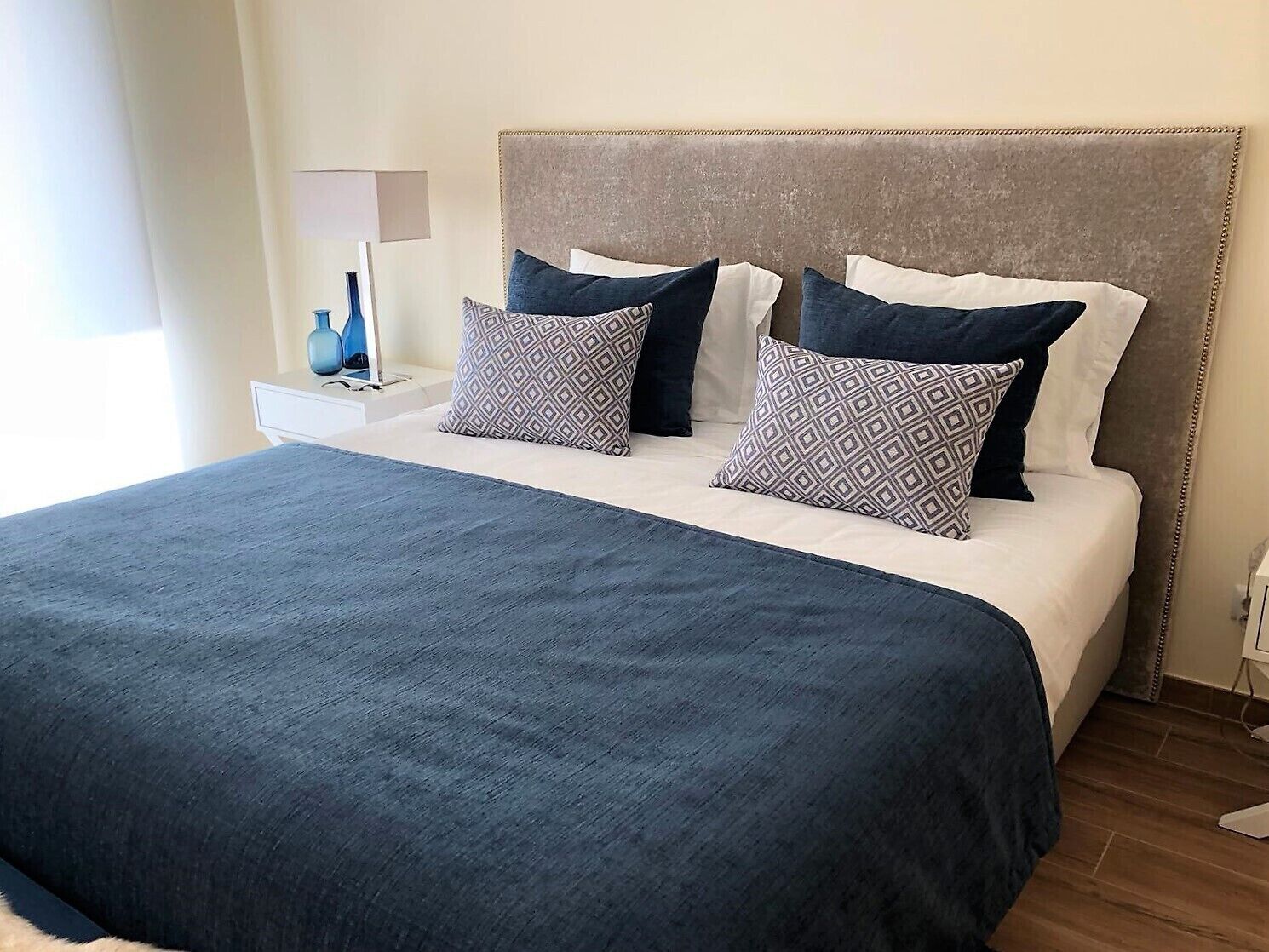 Un lit bien fait, recouvert d’une couvre-lit bleu foncé et agrémenté de deux coussins à motifs, une table de chevet avec une lampe et un luminaire fixé au mur.