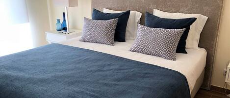 Un lit bien fait, recouvert d’une couvre-lit bleu foncé et agrémenté de deux coussins à motifs, une table de chevet avec une lampe et un luminaire fixé au mur.