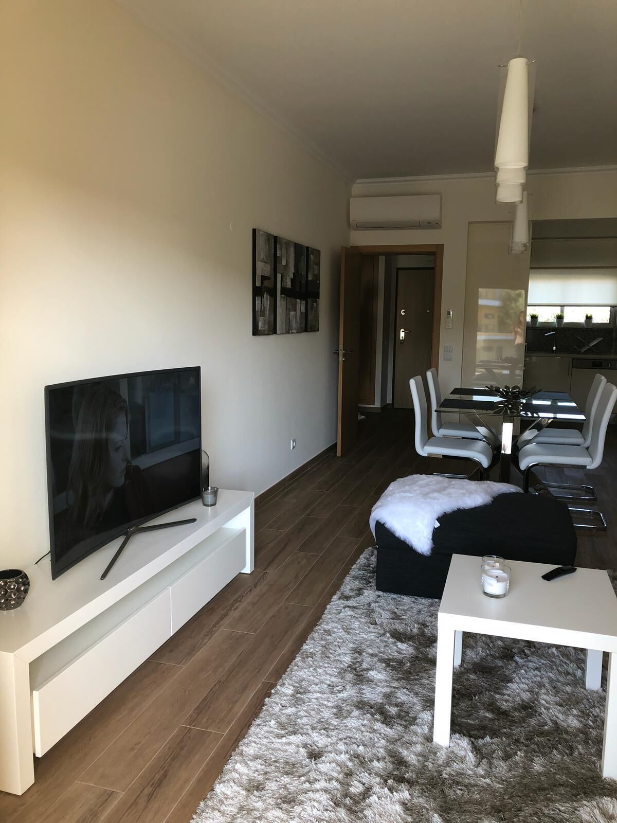 Ett modernt vardagsrum med en platt-TV, ett vitt soffbord, en mjuk matta och en matplats med ett bord och stolar.