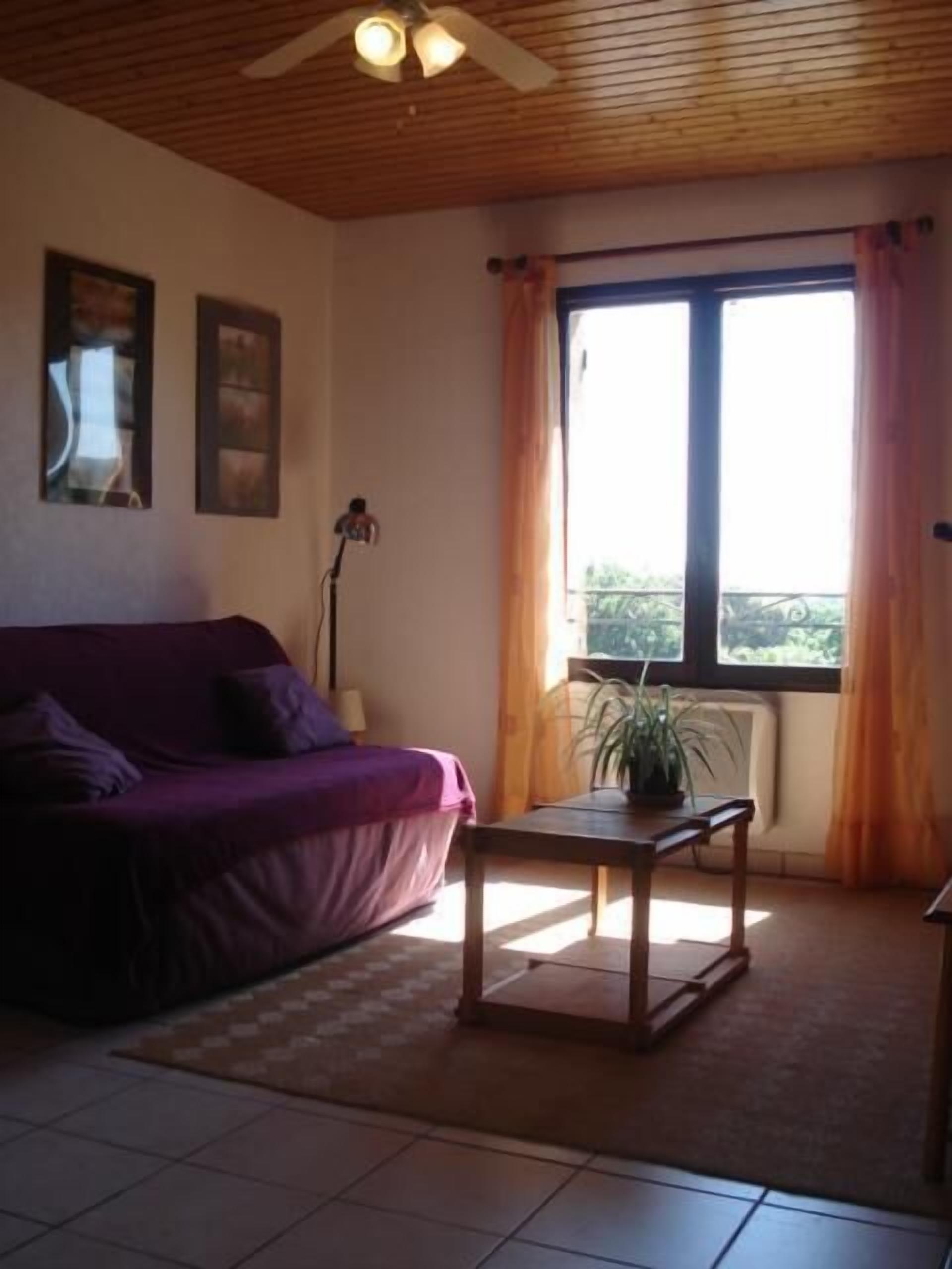 A room with a purple sofa, a wooden table, a ceiling fan, and a window with curtains.