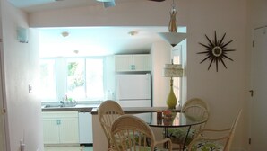 A dining area with a round glass table and four wicker chairs, a kitchen with white cabinets, and a decorative sunburst wall piece.