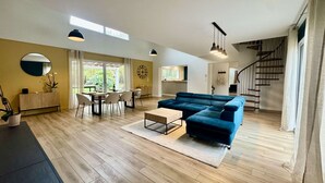 Amplia sala de estar con sofá azul, piso de madera, una mesa de comedor con sillas, una cocina al fondo y una escalera.
