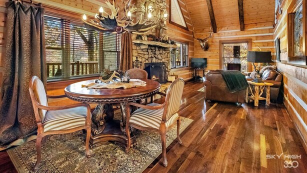 A rustic cabin interior with a round wooden dining table, striped chairs, a fireplace, and a stone wall. - Cinnamon Valley - The Elk Lodge (Eureka Springs)