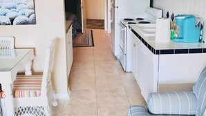 A compact kitchen with white cabinets, a stove, and a microwave. There is a dining area with a small table and a chair, and a sofa with striped cushions. A painting of a beach scene is visible on the wall. - 338 Ocean View!  Quiet Villa W/ Many Amenities 550 Sq FeetVRBO#394687 (Hilton Head Island)